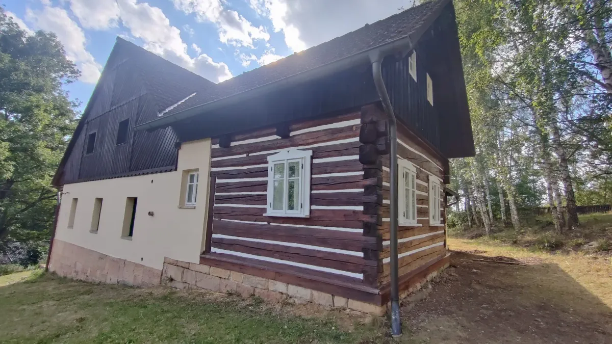 Stejný roh roubenky po nátěru, tmavé trámy a&nbsp;bílé spáry, vedle světlá zděná část